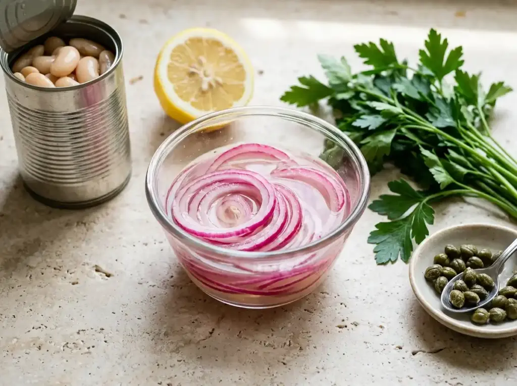 Sliced red onion soaking in lemon juice next to cannellini beans, parsley and capers